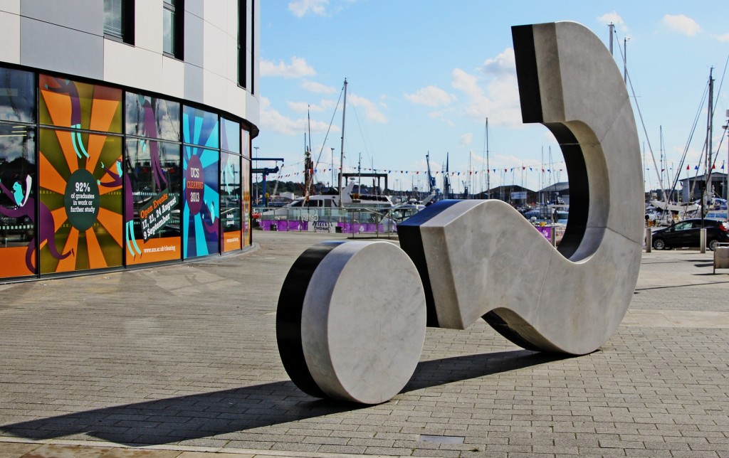 sculpture of a question mark on its side in front of masts in a harbour