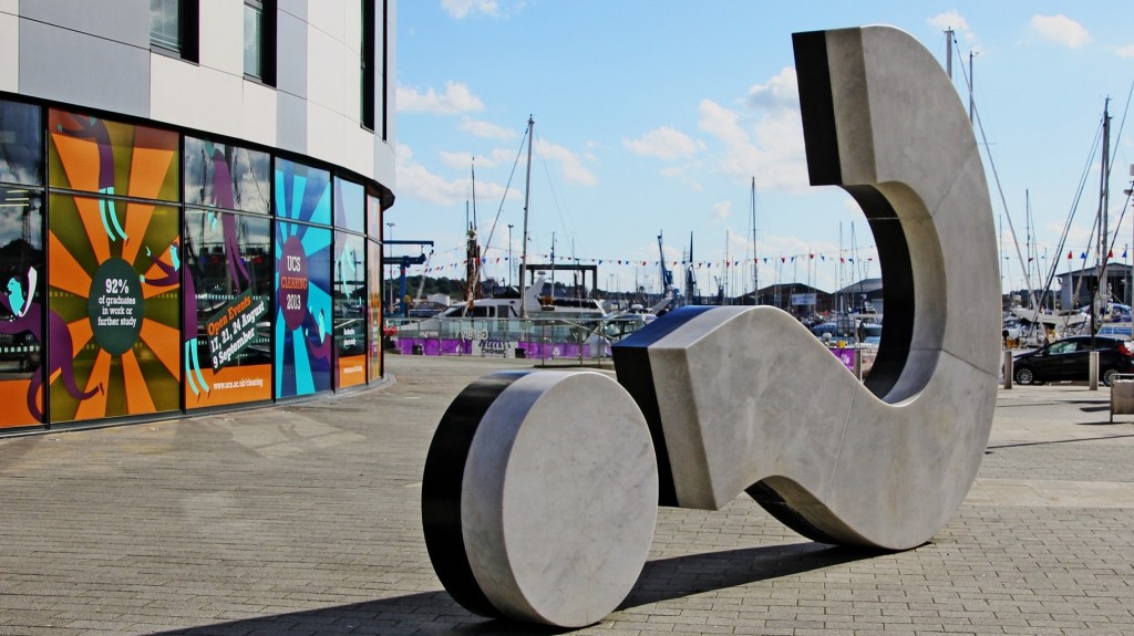 sculpture of a question mark on its side alongside the masts in a harbour