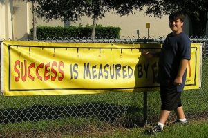 boy standing in front of a banner reading 'success is measured by...'The final word is obscured