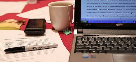 paper and pen alongside an online article on a laptop