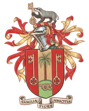 A coat of arms, mostly in red and gold. It features a badger, gold keys, a palm tree, and an artillery gun. The motto is 'Family, faith, friendship' (in Latin)