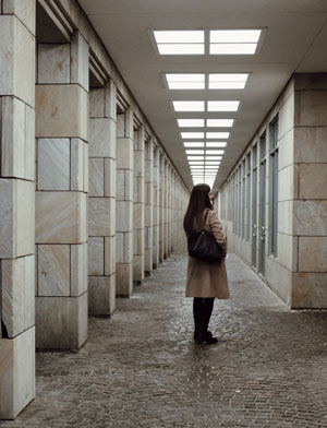 A woman stands alone in the middle of a corridor with many stone pillars