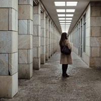 A woman stands alone in the middle of a corridor with many stone pillars