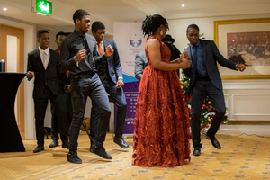 A Black woman wearing a beautiful red evening dress is dancing with a group of five Black teenage boys who are smartly dressed in suits and ties.