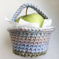 A small crochet basket in shades of grey and blue yarn, just big enough to hold a green apple surrounded by some white paper