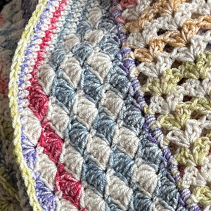 Detail of a crochet blanket. The border is in a shell-shaped stich, with the rows in a v-shaped stitch. Muted rainbow colours alternating with cream.