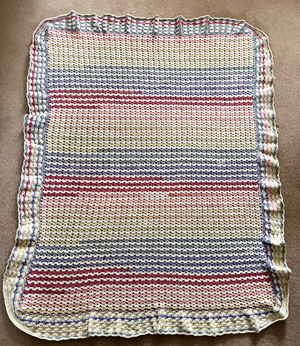 A crochet blanket. The centre is horizontal stripes in muted rainbow colours. The border is ruffled.