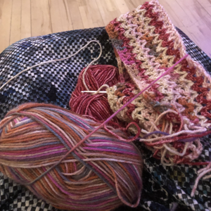 A crochet project, with a yarn in shades of rust and cream that is being made into a small blanket that has just started. Glimpse of a blue patterned dress and a wooden floor.