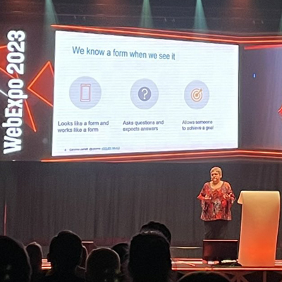 Presenting at the WebExpo conference. Caroline is wearing a red top and black trousers, standing next to a beige lectern and in front of a giant screen with one of the slides from a talk
