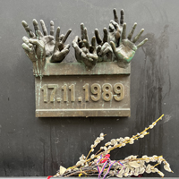 The monument to the Velvet Revolution. It's a small plaque on a wall. The top of the bronze plaque has life-sided sculptures of 8 hands, above the date 17.11.1989. Below the plaque, someone has left a branch of dried flowers.
