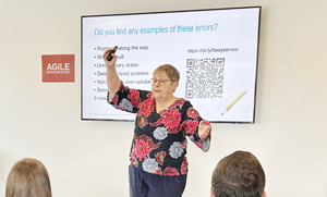 Caroline Jarrett talking at a conference. She is wearing a black top with large ping and blue flowers on it, and waving her ams in front of a slide asking people to chat about errors.