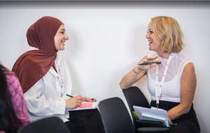 A woman wearing a brown hijab is chatting to a woman with blonde hair. They both have white tops and are holding pens and sticky notes.