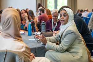 People working at round tables, including a woman wearing hijab talking to someone else wearing hijab